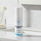 A white cylindrical bottle of Balanced Bayou Niacinamide Serum stands on a white marble countertop in a bathroom setting with a sink and vase in the background.