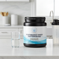 A tub of Balanced Basics Performance Whey Protein Blend in Chocolate flavor sits on a white kitchen counter next to a glass of water and a shaker bottle.
