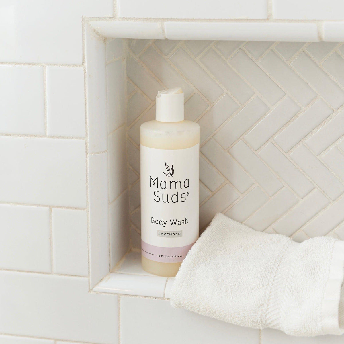 Mama Suds body wash bottle on a tiled shower wall with a towel.