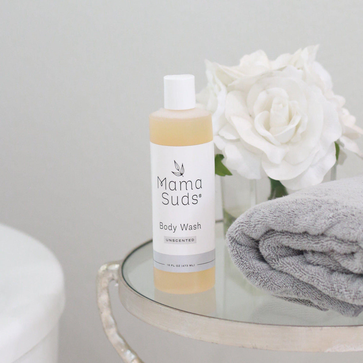 Mama Suds body wash bottle on a glass table with a gray towel and white flowers.