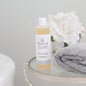 Mama Suds body wash bottle on a glass table with a gray towel and white flowers.