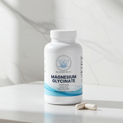 A white bottle of Balanced Bayou Magnesium Glycinate capsules sits on a light marble surface with two capsules placed in front. The background is a soft, out-of-focus white marble setting.