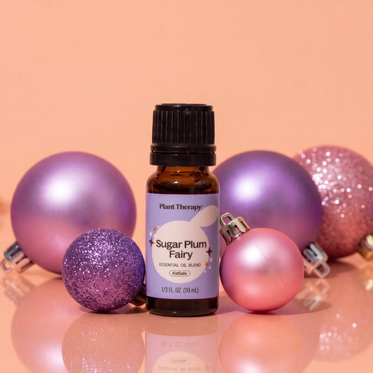 A small amber glass bottle labeled "Sugar Plum Fairy" by Plant Therapy sits on a reflective surface, surrounded by shiny and glittery purple and pink ornaments against a peach-pink background.