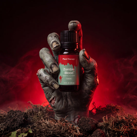 A small amber bottle labeled Plant Therapy Zombie Blood essential oil is held in a zombie-like gray hand rising from dark soil, with red mist and a black background.