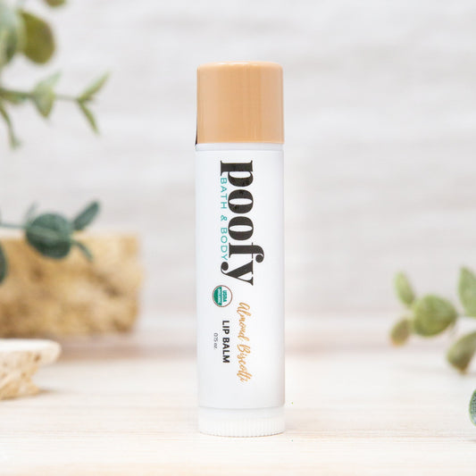 A tube of Poofy Natural Balm almond butter lip balm stands upright on a light wooden surface with soft green leaves blurred in the background.