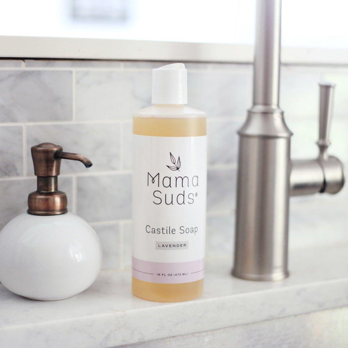 Mama Suds Castile Soap bottle on a kitchen counter with a soap dispenser and faucet in the background.