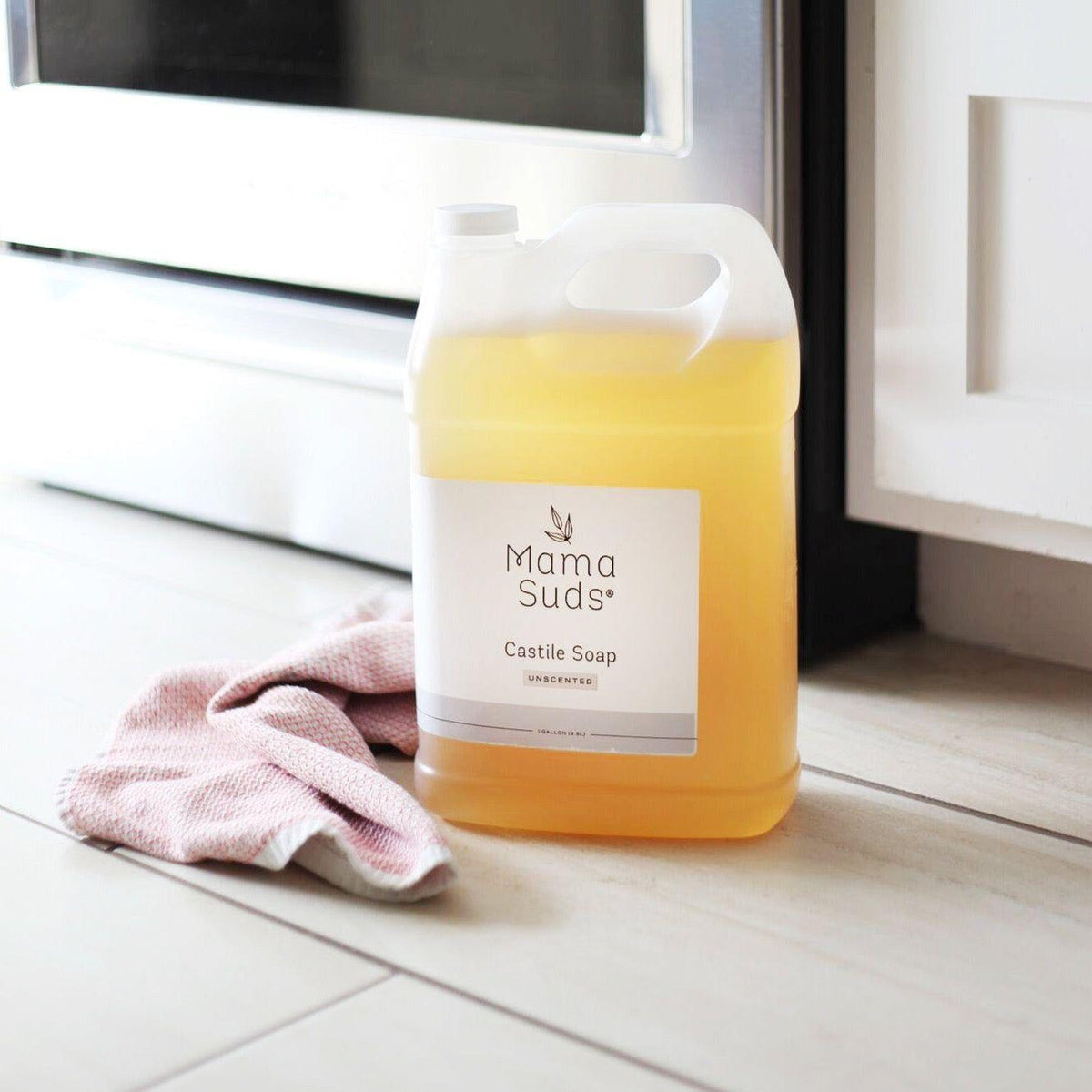 A large plastic jug of Mama Suds Castile Soap Undiluted with a white label, filled with pale yellow liquid, sits on a light wood floor next to a pink cloth near a white appliance.