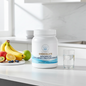 A white tub of Chocolate Advanced 100% Whey Protein Isolate by Pure Encapsulations sits on a white kitchen countertop, styled with fresh fruit and a glass of water in a bright, modern kitchen setting.