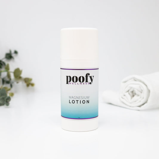 A white cylindrical bottle of Poofy Wellness Magnesium Lotion with a purple and blue gradient label, set against a clean white background with soft-focus decorative greenery and a rolled white towel.