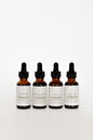 Four amber glass dropper bottles with black caps and white labels, arranged in a row on a clean white background. The labels show different scent names including Saffron, Mullein, Lunna, and Oil of Oregano.